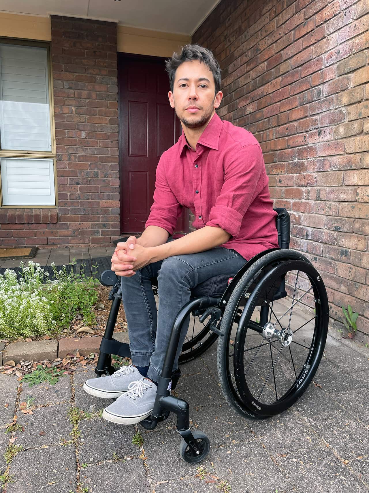 A man with short brown hair sits in a sleek black manual wheelchair, looking directly at the camera with a neutral expression. He is wearing a long-sleeved dark red button-up shirt and dark grey jeans, positioned on a paved walkway in front of a brick house.