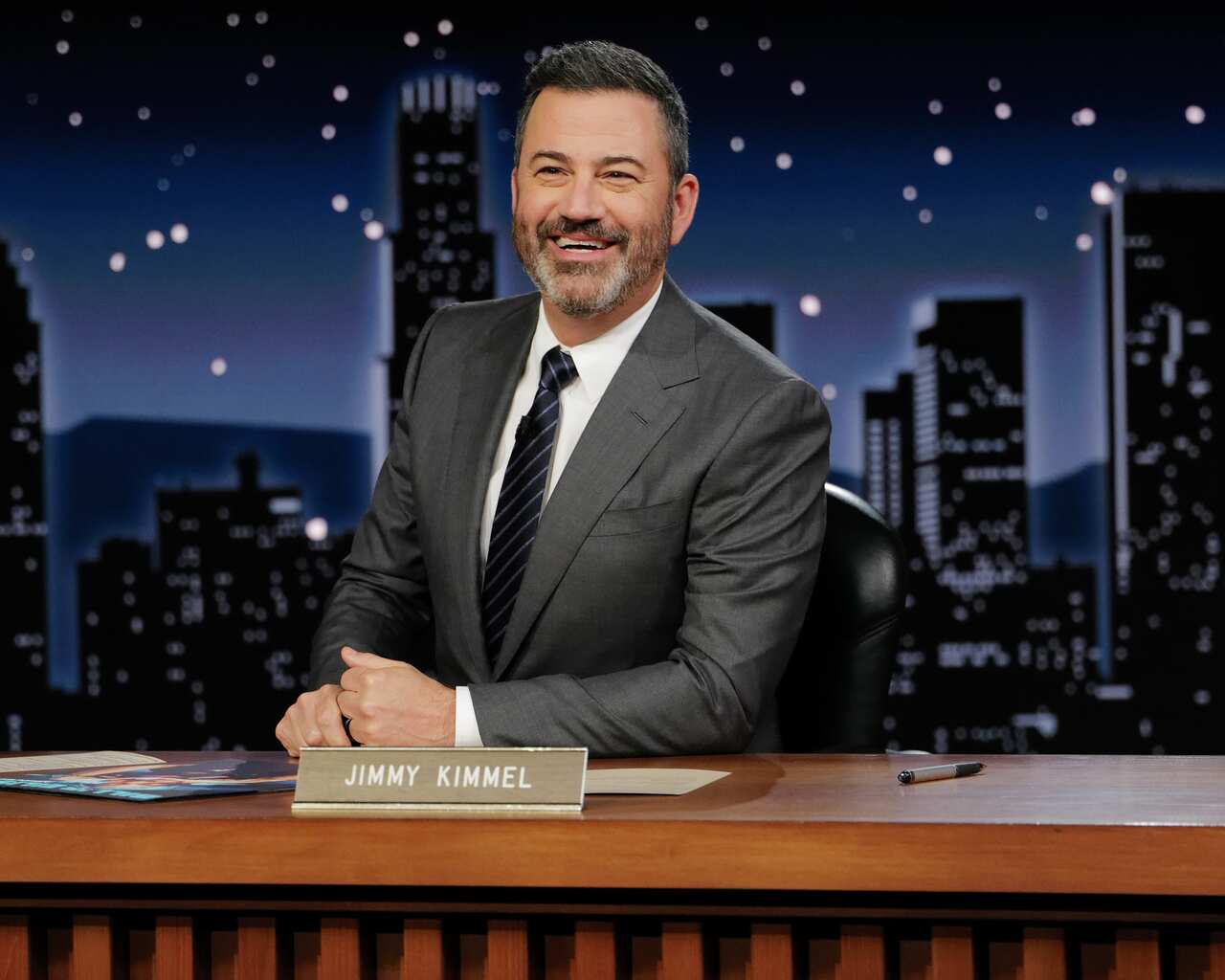 Jimmy Kimmel smiles as he is seated at a desk.