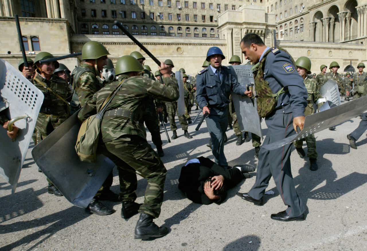 Police and soldiers stand over a man while swinging batons.
