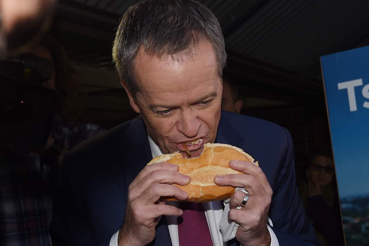 Bill Shorten eats a sausage in a roll. He is holding the roll sideways to his mouth