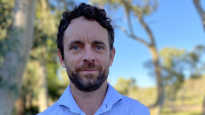 A headshot of a man in a blue shirt with trees in the background