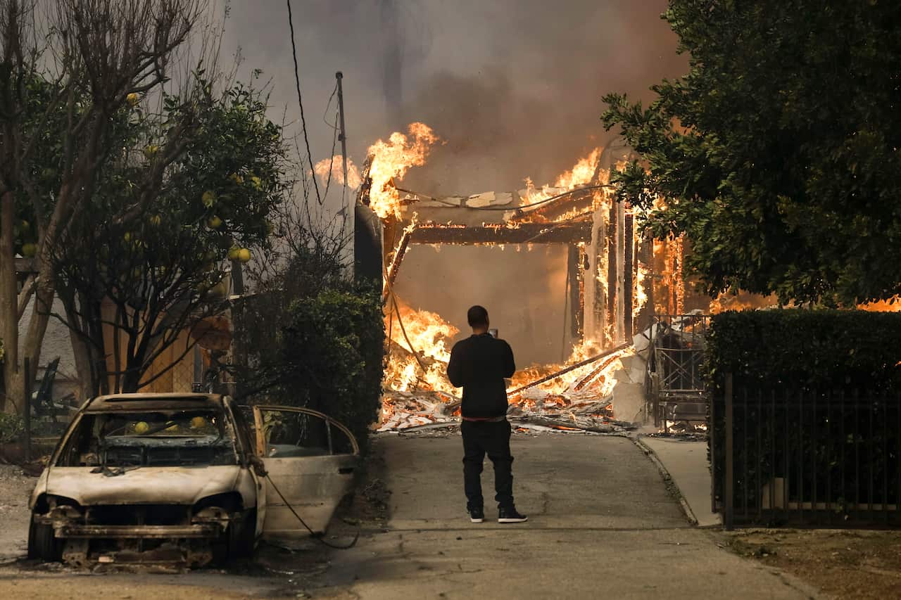 A person watches as a house burns, with a charred car beside him.