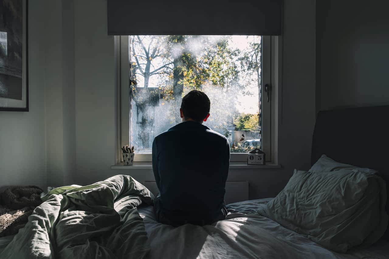 Rear view of an unrecognisable man sitting on his bed looking out of the window - negative emotion