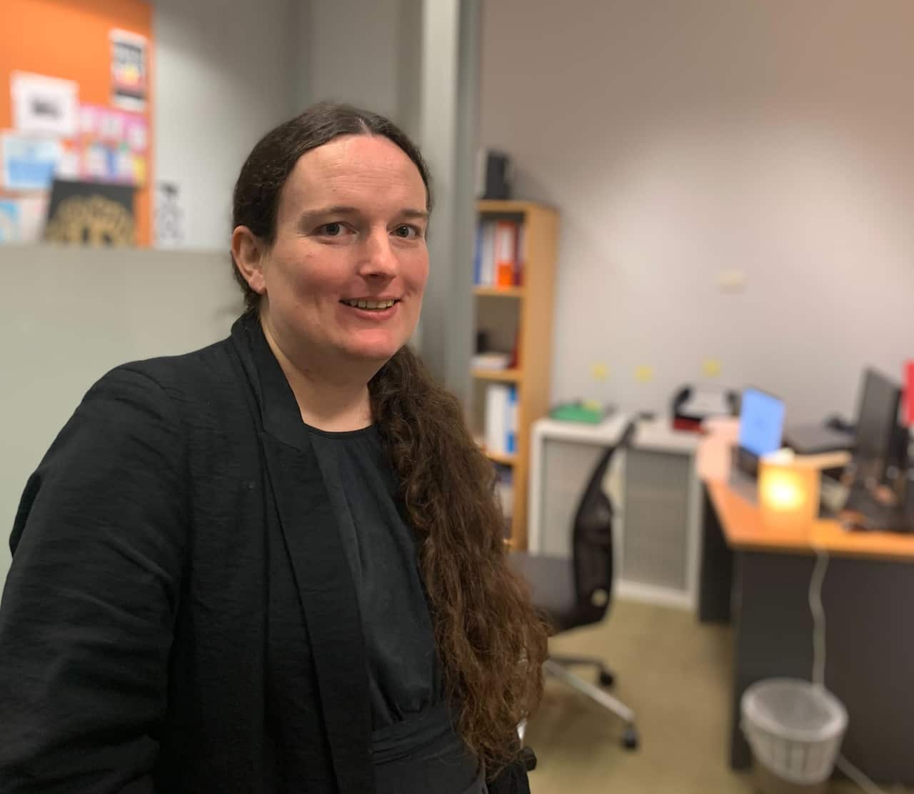 A smiling woman with long brown hair, wearing a dark top and jacket, poses for a photo in an office. 