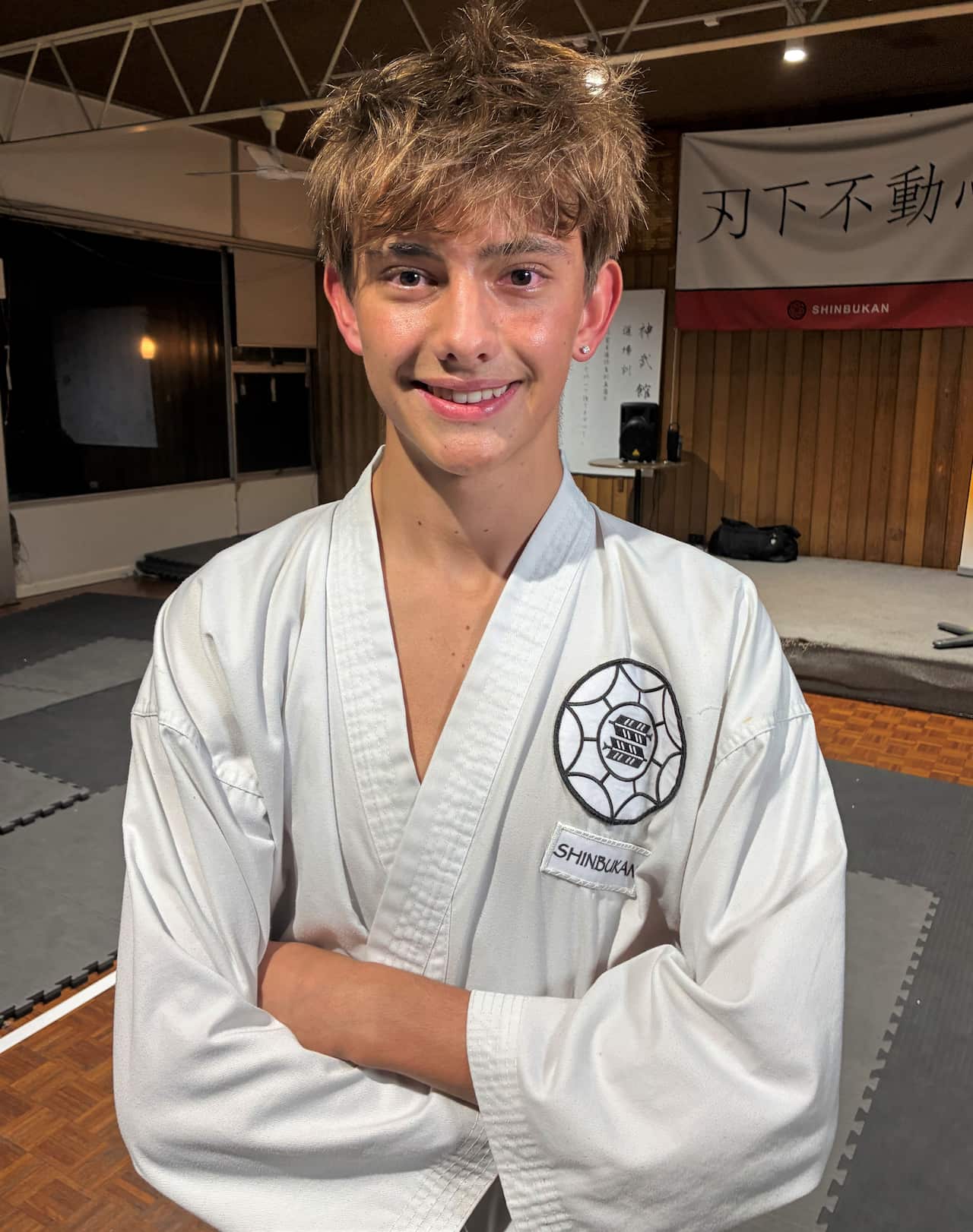 A man with light brown hair stands with folded arms wearing a white karate jacket.