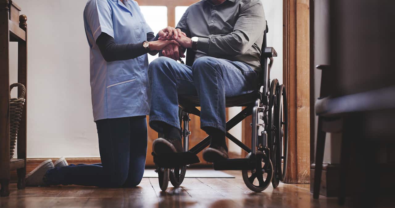 A carer kneels beside a person in a wheelchair, no faces are shown.