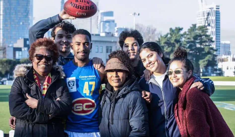 Hewago Oea wearing an Aussie rules jumper poses for a photograph with a small group of people