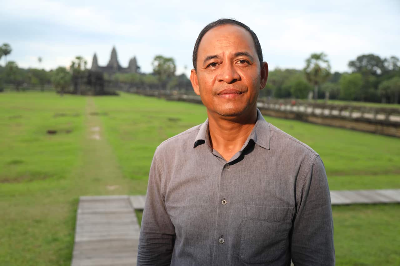 A middle-aged Cambodian man in a grey shirt poses for a photo with Angkor Wat temples in the background 