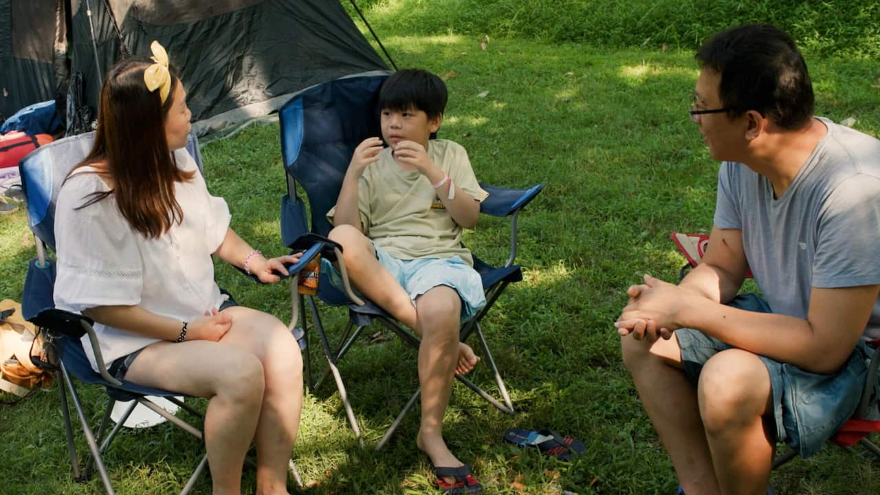 A family of three are sitting in camping chairs. A woman and a man look at their son in the middle who is gesturing in sign language 