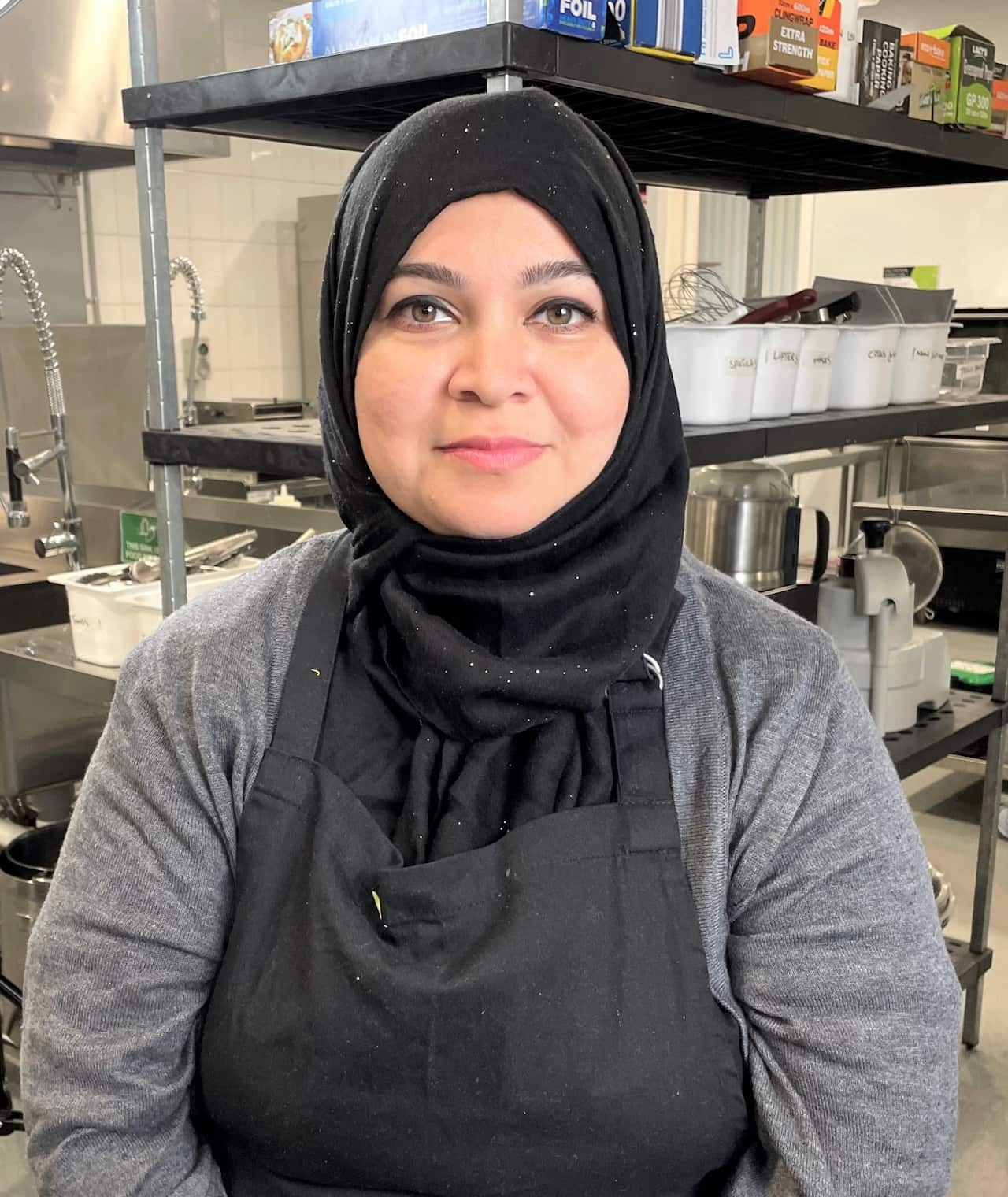 A woman in a grey shirt sitting in front of kitchen equipment.