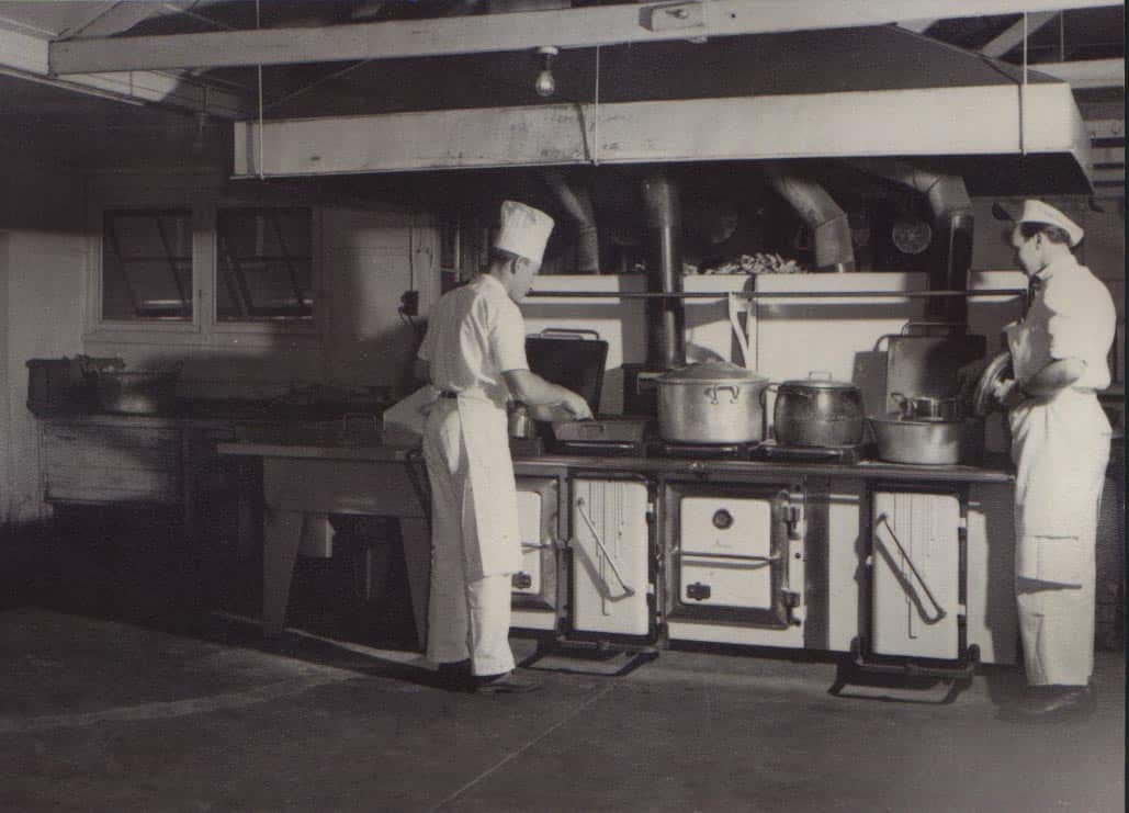 A black and white photo of two men in chef's hats cooking in an old-fashioned kitchen