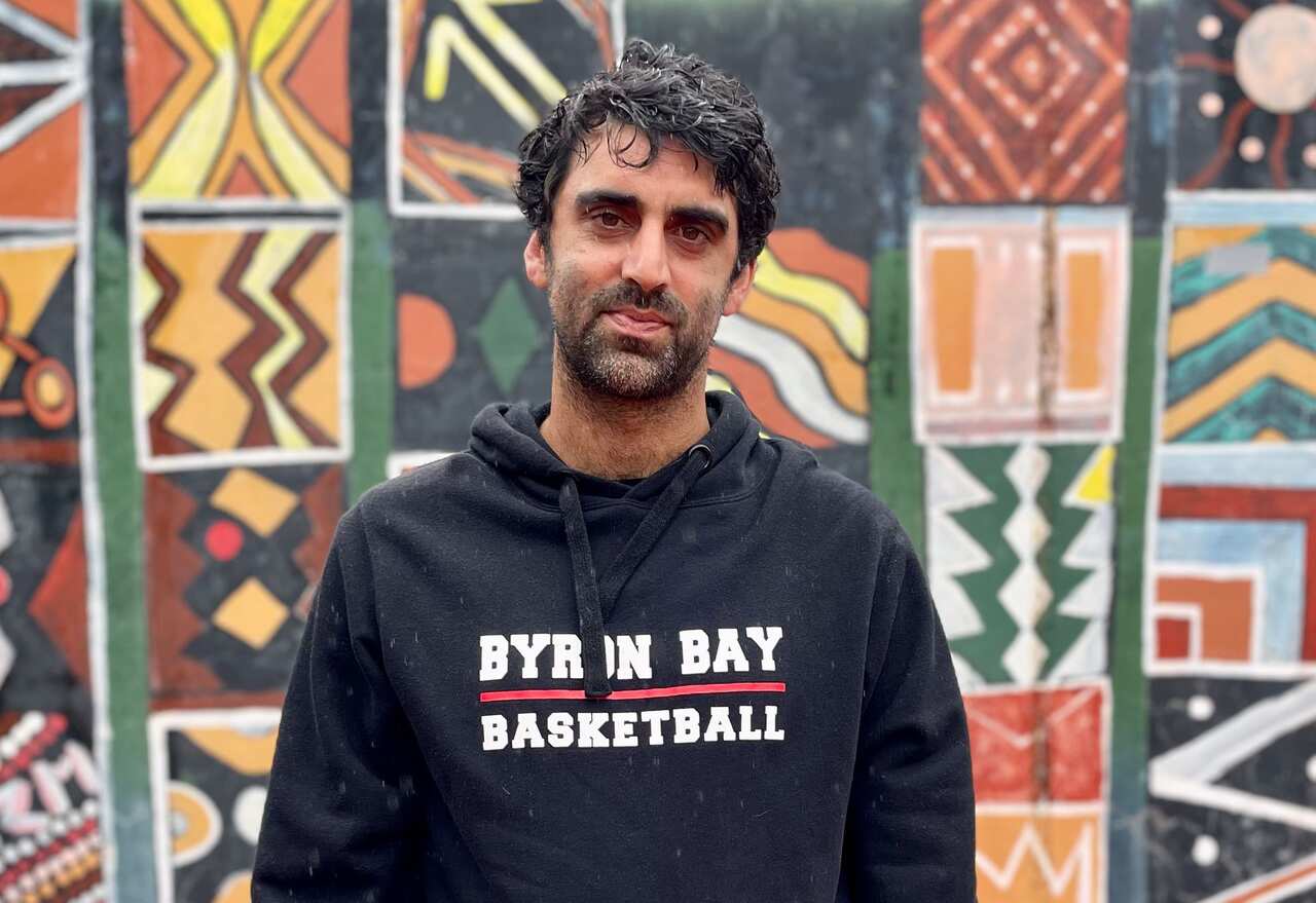 A man in a black hoodie that reads "Byron Bay Basketball" standing in front of a colourful wall.