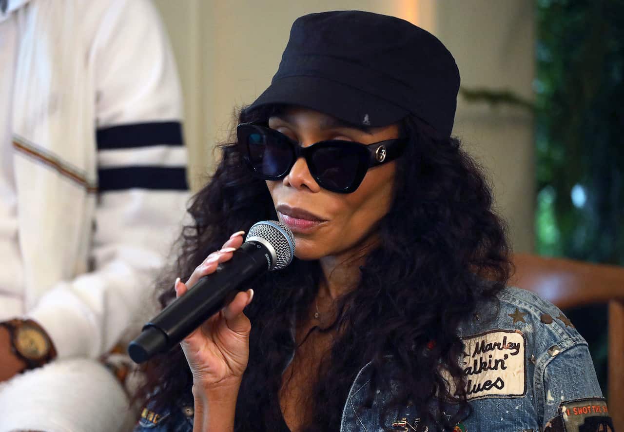 A woman wearing black sunglasses, a black cap and a blue denim jacket, holding a microphone.