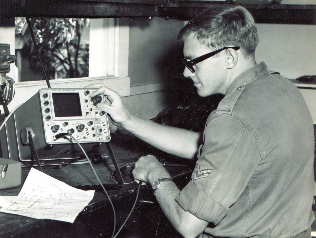 A black and white image of a man wearing an army uniform sitting at a desk and turning a dial on a piece of electronic equipment