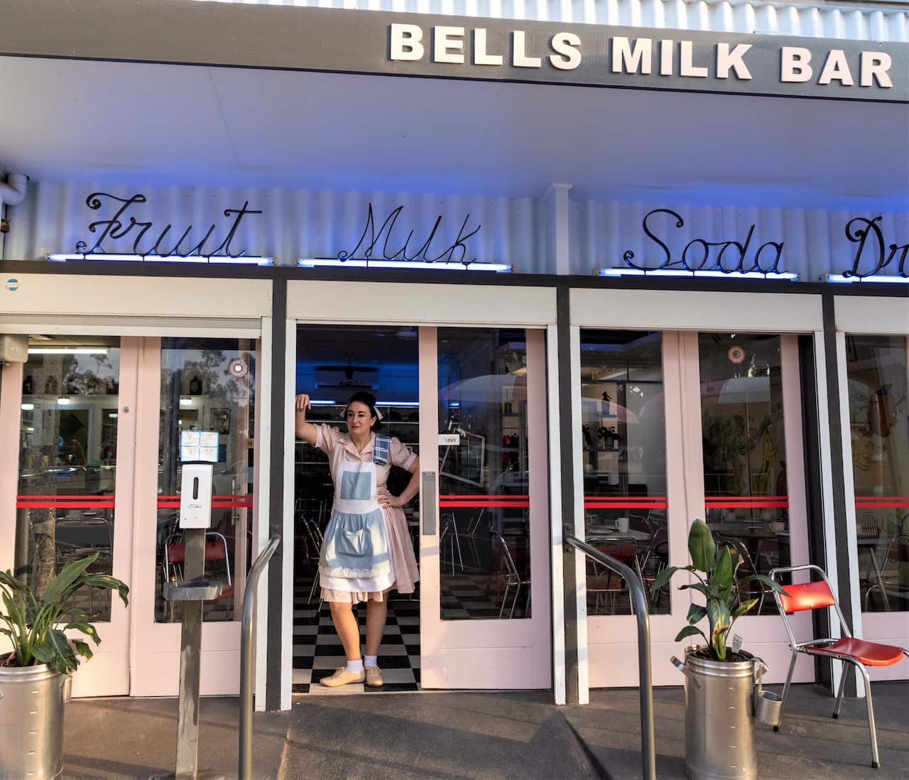 A woman in a waitress uniform leans against the door frame of a milk bar painted in vintage colours. 