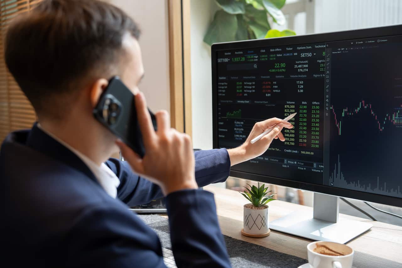 A young man wearing a suit points to his computer screen where numbers are displayed, as he talks on the phone