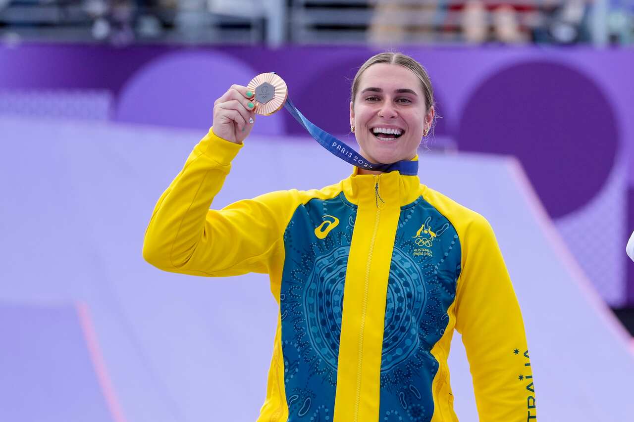 A woman wearing a yellow and green tracksuit holding up a bronze medal with her right hand