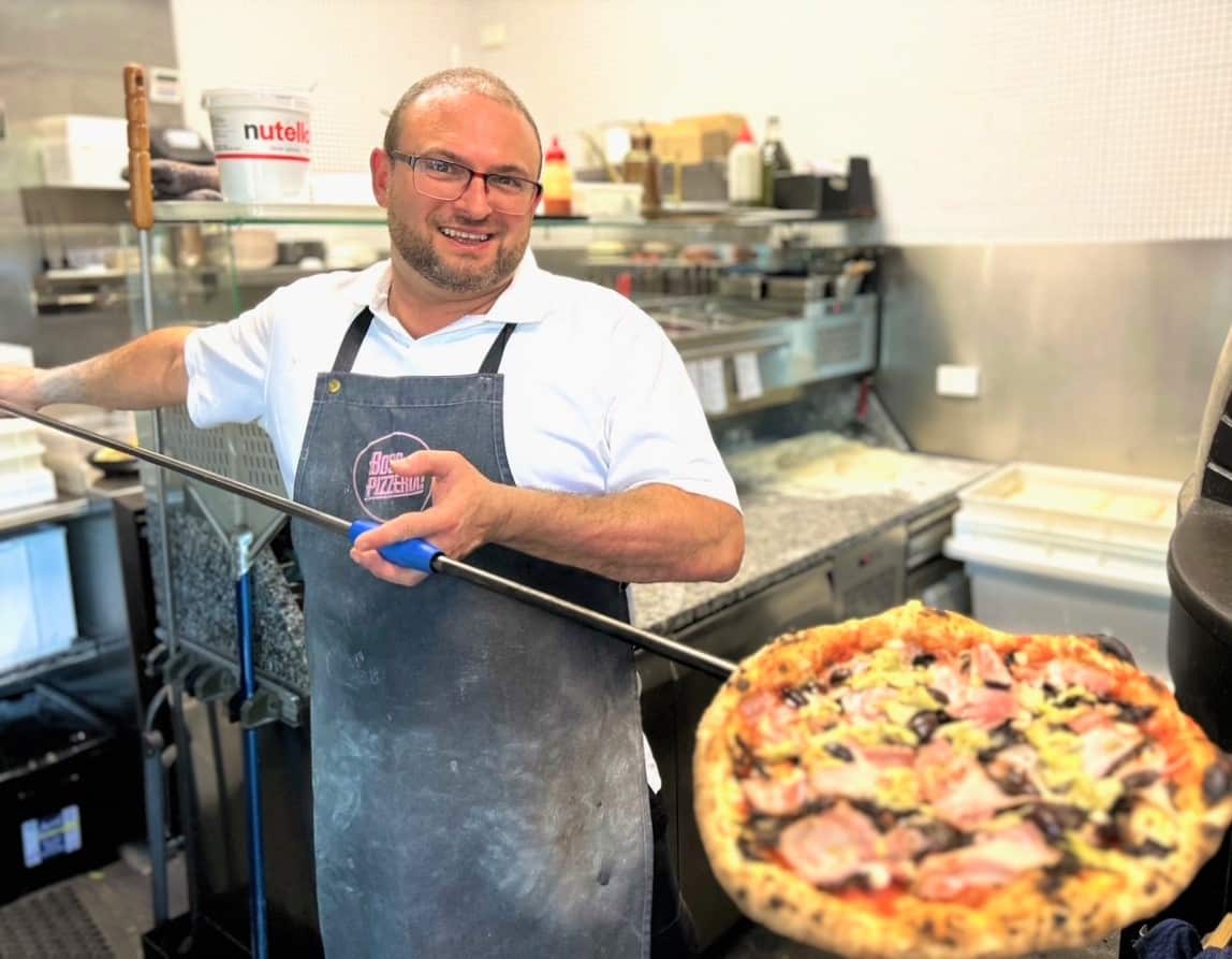 A man in a white apron stands lfting a pizza from a wood-fired oven.