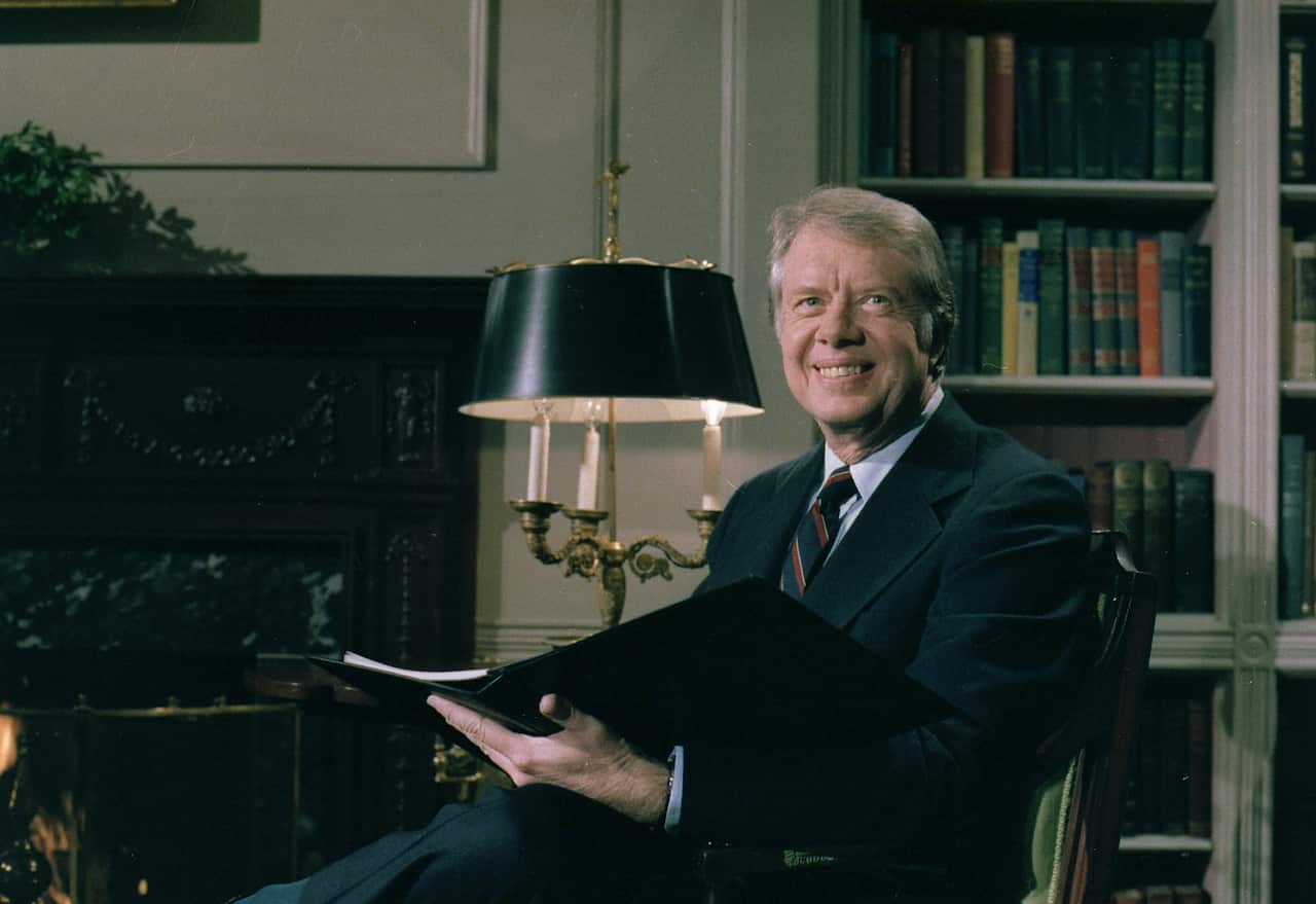 A man wearing a suit and tie smiles as he holds a file