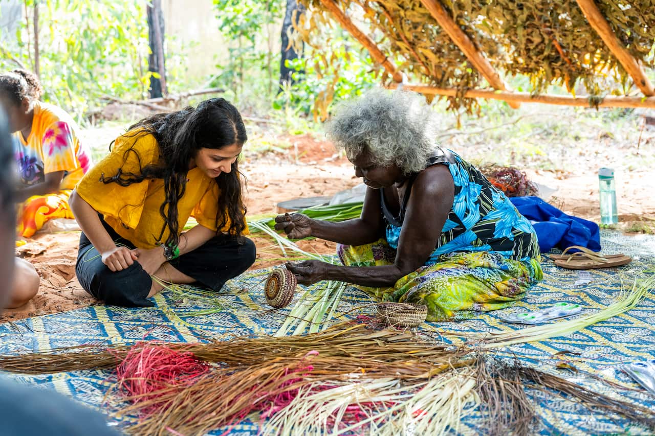 Two women sitting on the ground. One is showing the other how to weave.
