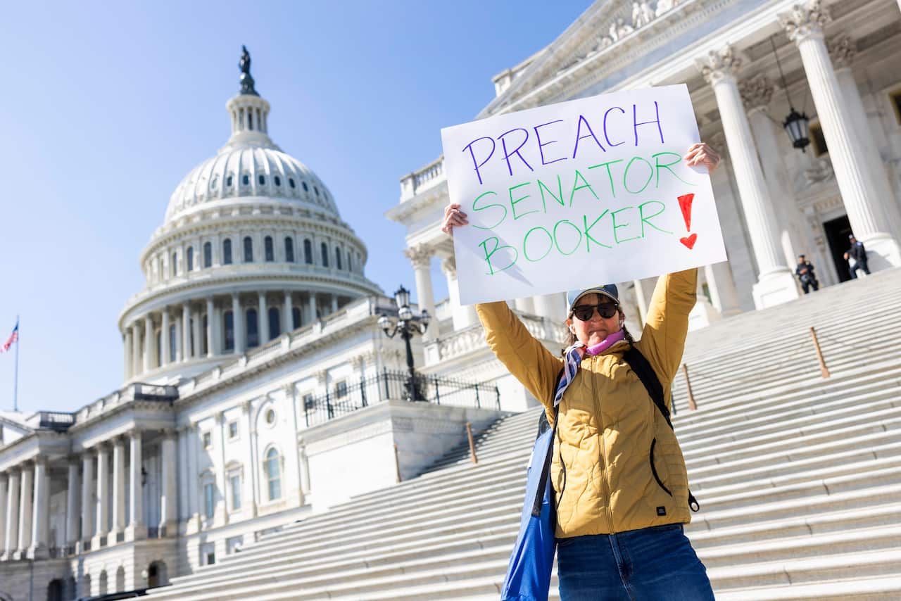 A woman standing on the steps of a building holds up a hand-written sign that reads "Preach Senator Booker!"