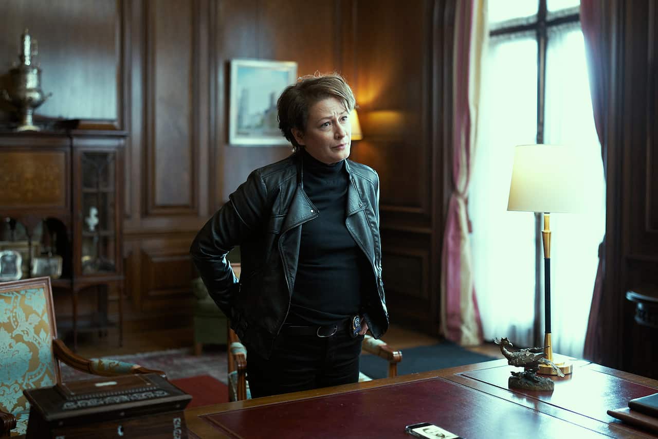 A woman in a leather jacket stands at a desk in a dark-panelled room.