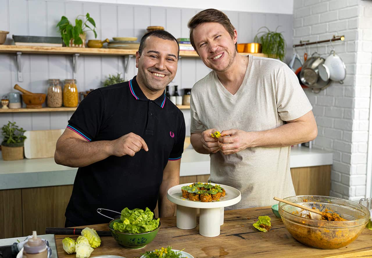 Two men stand behind a ktichen bench, with food in bowls and on a stand in front of them. They are both smiling. 