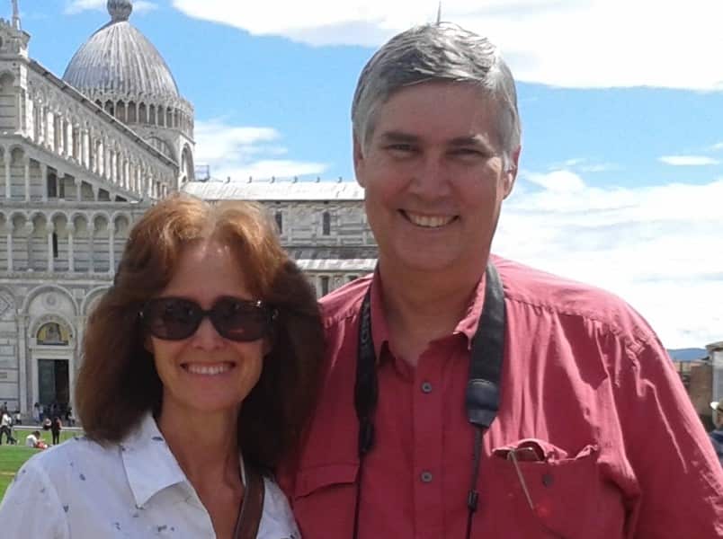 A man in a red shirt stands next to his female partner, who is wearing a white shirt and sunglasses. They are both smiling.