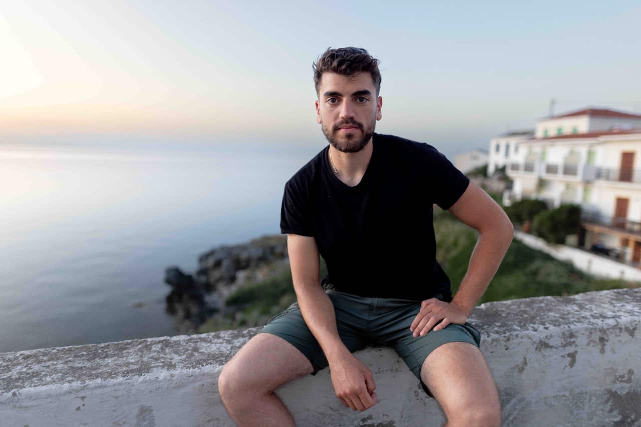 A young bearded man wearing a black t-shirt and grey shorts sits on a low wall. Behind him is the sea at sunset, and white buildings.