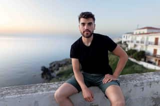 A young bearded man wearing a black t-shirt and grey shorts sits on a low wall. Behind him is the sea at sunset, and white buildings.