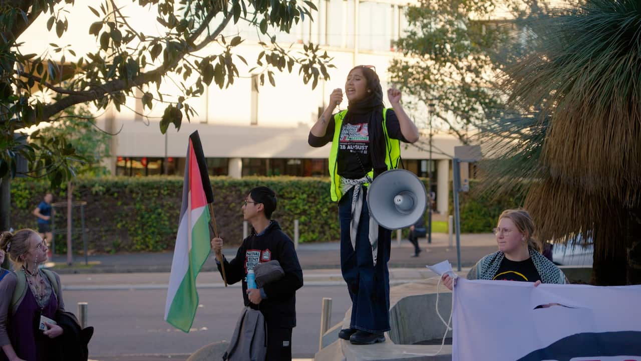 A woman wearing a high-vis vest speaks into a megaphone while standing on a table outside. A man stands in front of her holding a Palestinian flag and two othe people look on.