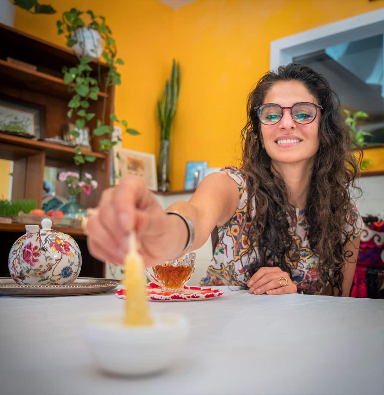 A woman wearing glasses and a floral dress sits at a table with a teapot.