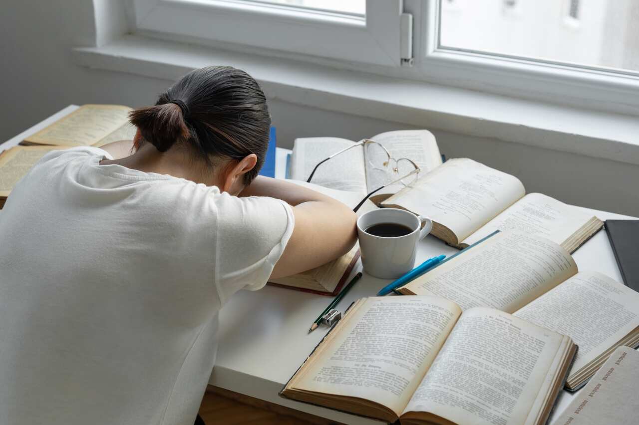 A student with their head down on the desk, surrounded by books and a cup of coffee.