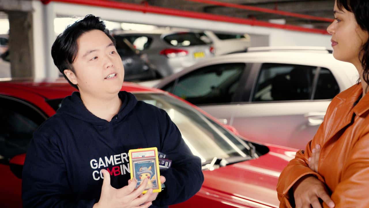 A man in a dark hoodie holding a Pokemon card in a plastic case looking at a woman in an orange jacket.  They are standing in an underground car park with cars parked