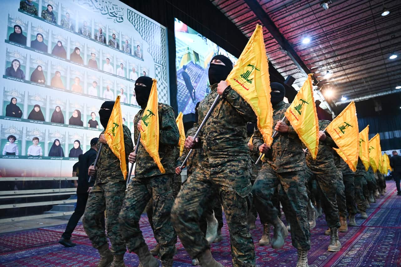 A group of men marching in military uniforms and wearing black face coverings while holding yellow flags