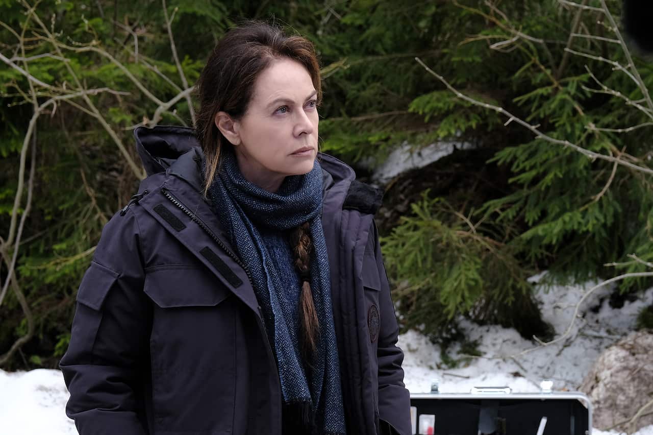 A woman in a coat and scarf stands outdoors in the snow, with trees behind her.