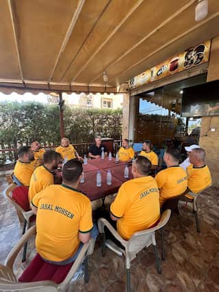 The Australian ambassador to Lebanon sits around a table with Jabal Mohsen locals wearing green and gold shirts, they each have a bottle of water on the table in front of them.