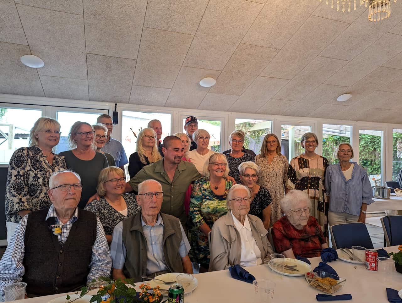 A large group of around 20 people ranging from middle aged to very old, posing for a photo around a table.