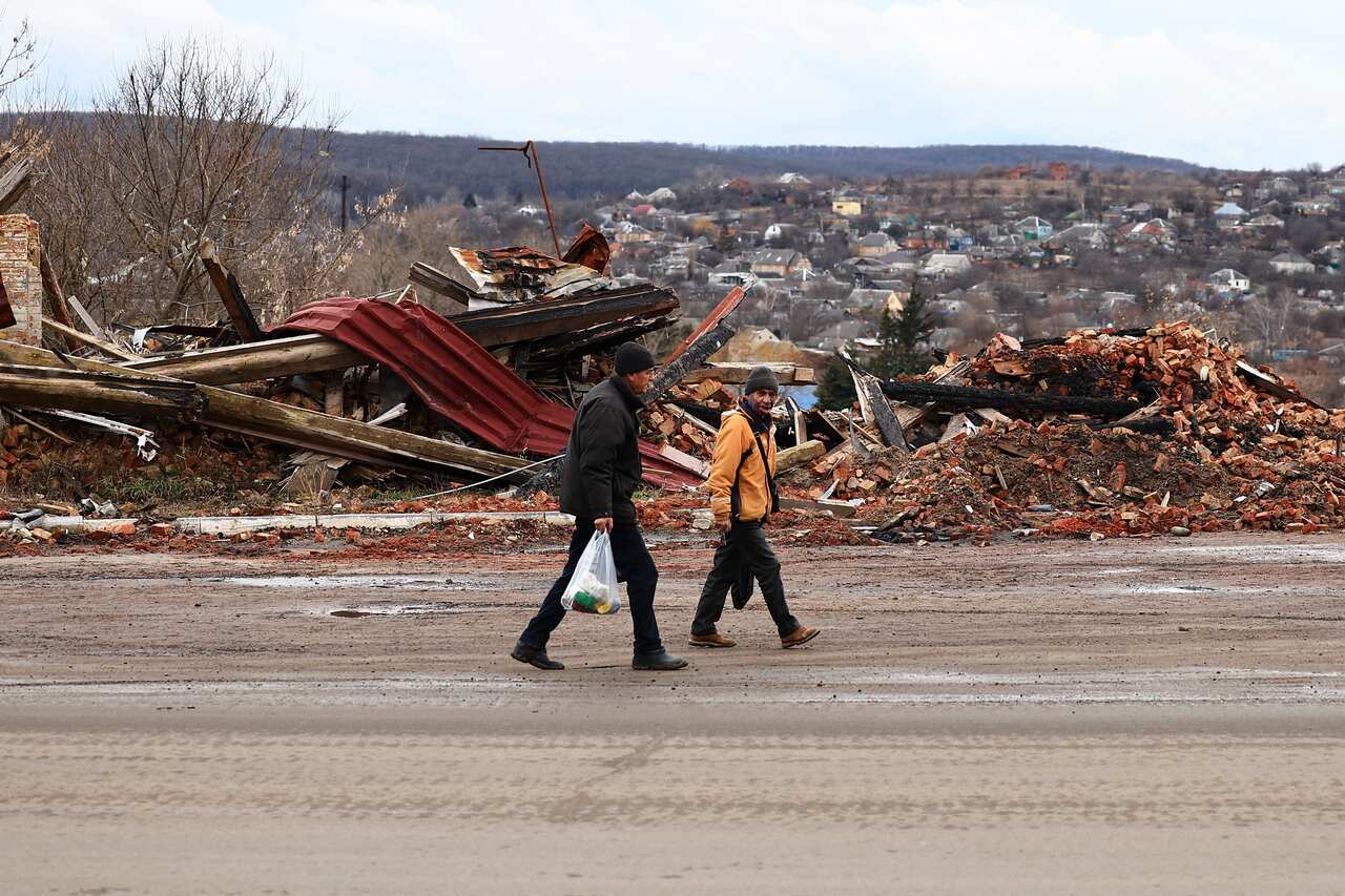 Two people walking along a road with destroyed buildings.