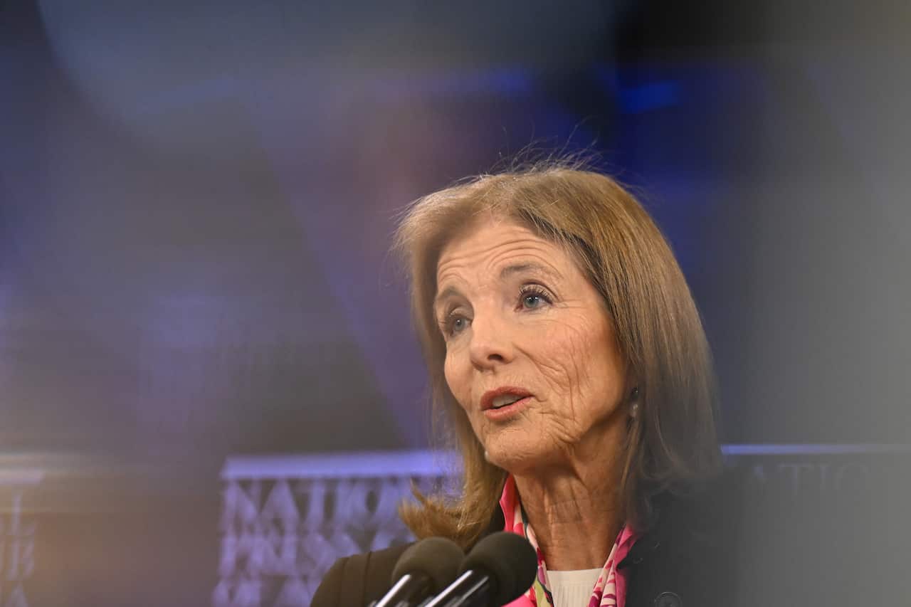 A woman in a blazer is standing in front of a blue wall, speaking into microphones.