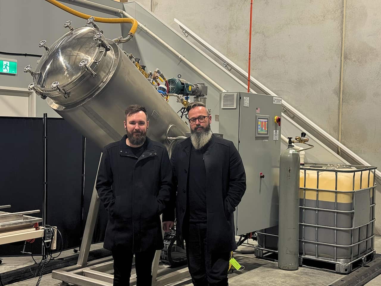 Two men in black coats stand in front of a stainless steel machine.