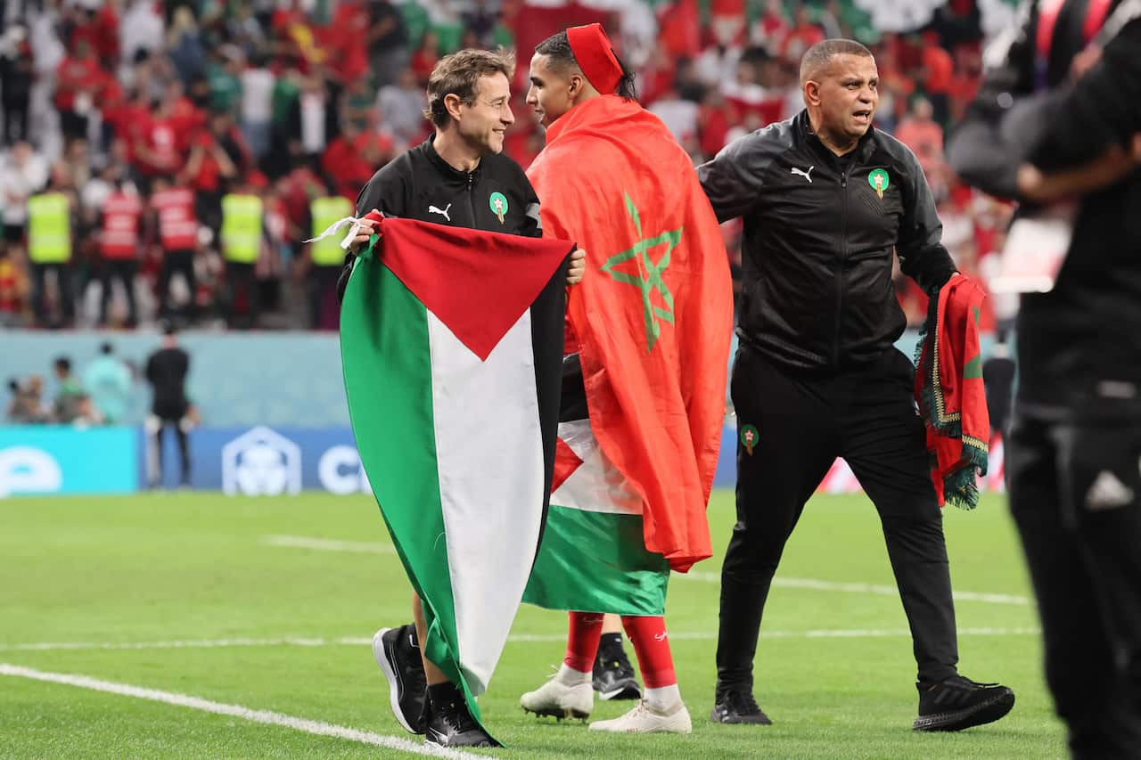 Man holds a Palestinian flag, and the other carries both a Moroccan and Palestinian flag on a field.