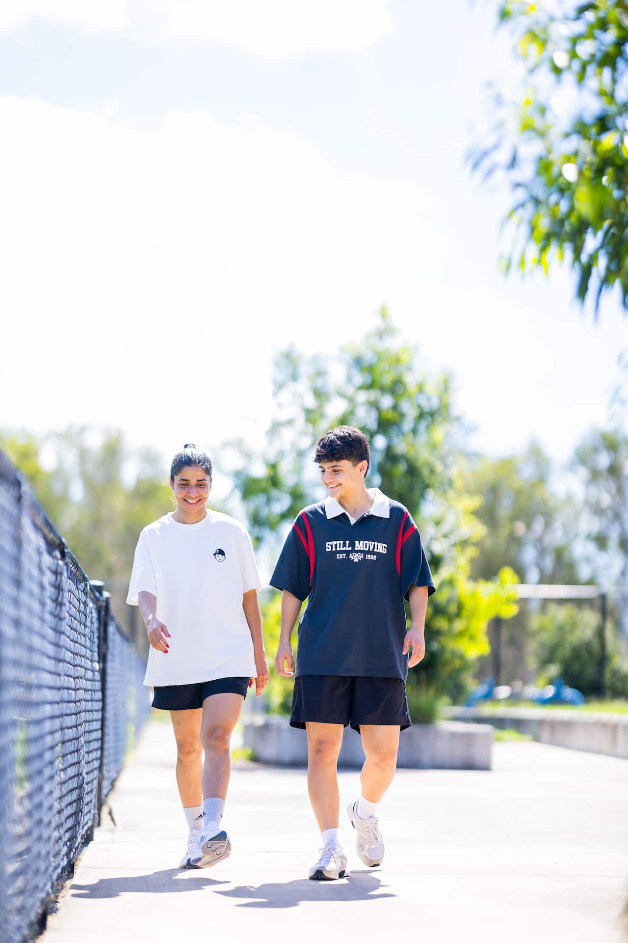 Two young women wearing shorts and short shirts walk together outside and smile.