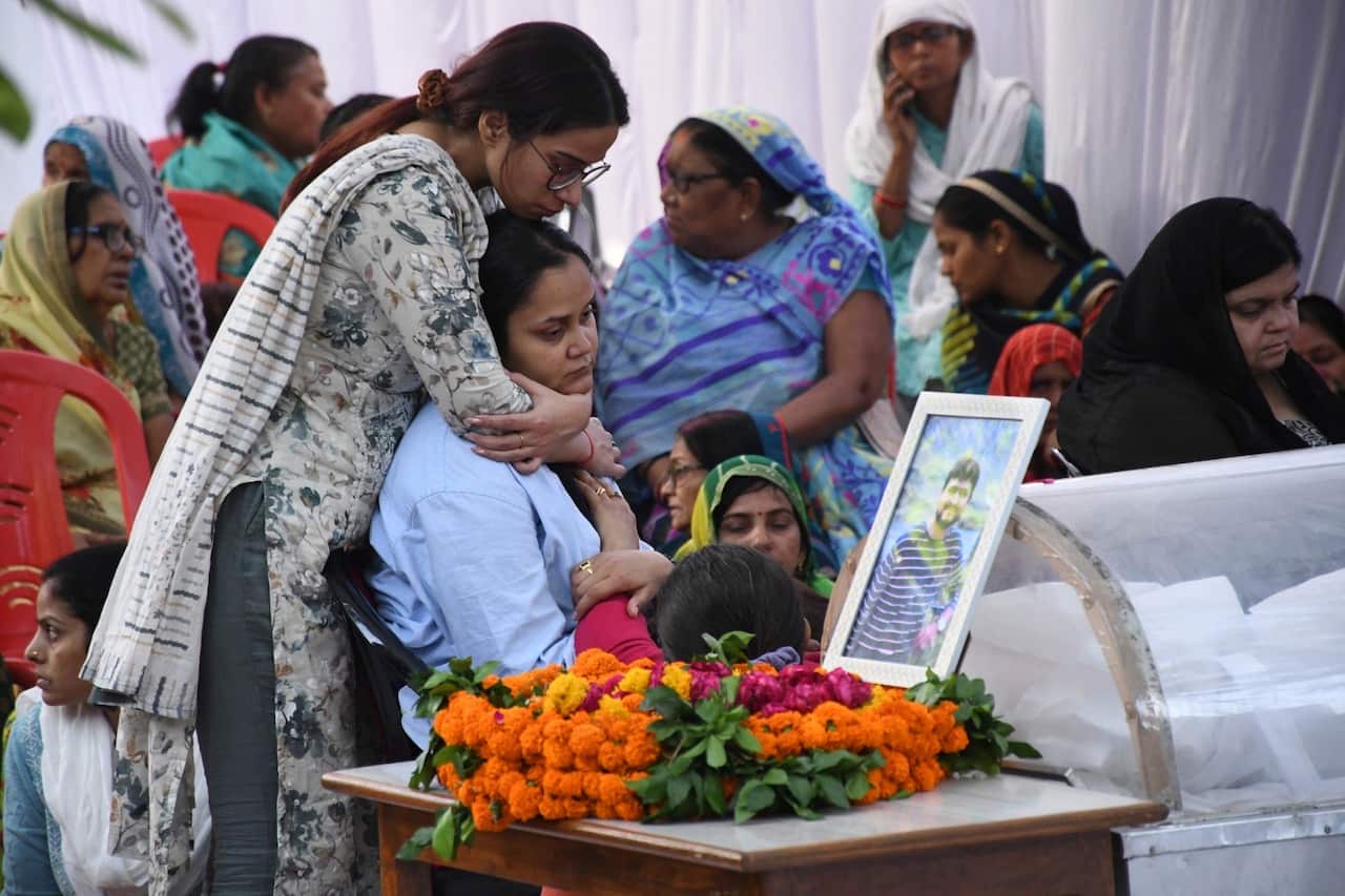 A group of women hold onto one another in front of a casket with flowers and a photograph.