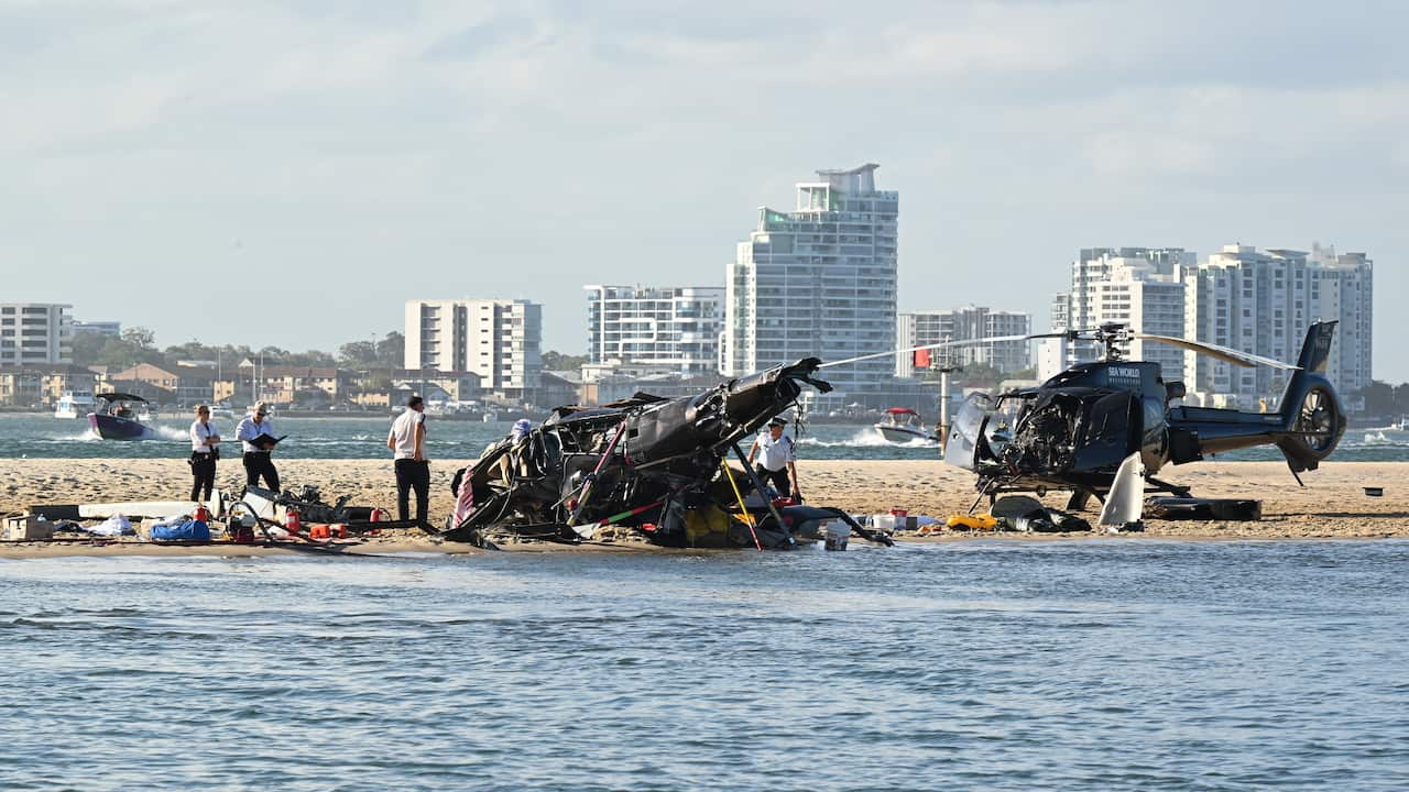 Two destroyed helicopters on a sandbar with people standing near them.