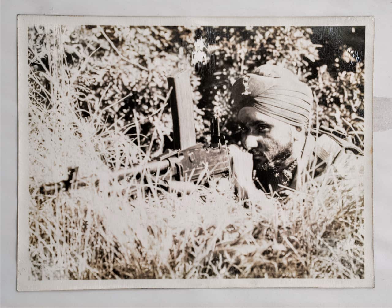 A black-and-white photo of a man wearing a turban crouches behind a hillock, pointing a military weapon.