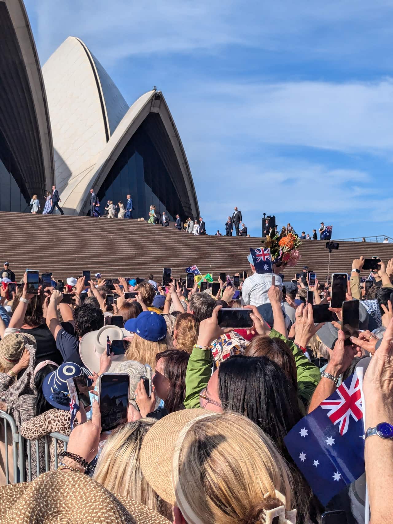 A crowd of people holding their phones in the air, at the steps of Sydney's Opera House