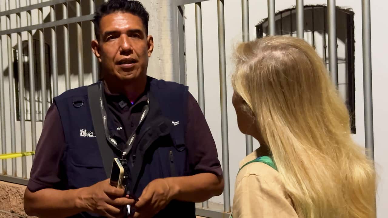 Two people stand next to a metal fence. On the left is a man wearing a dark vest holding a smartphone and appearing to be speaking, while a woman with long blonde hair listens facing away from the camera.