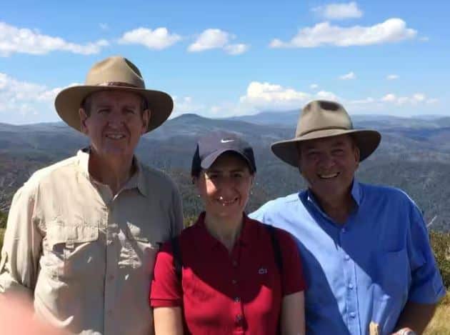 A photo of Barry O'Farrell smiling on the left, Gladys Berejiklian in the centre smiling wearing a hat, and Daryl Maguire wearing a hat on the left with his arm around her and smiling. The backdrop is mountain ranges. 
