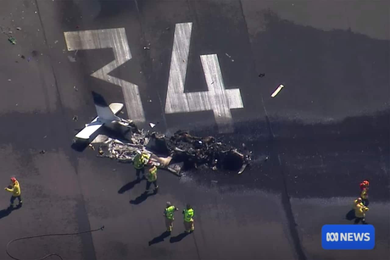 An aerial image of a burnt plane wreckage sitting on an airport tarmac. Emergency personnel surround the wreckage.  